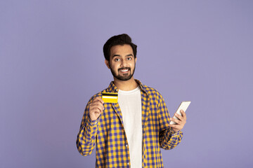 Online shopping. Cheerful Indian man holding mobile phone and credit card on lilac background