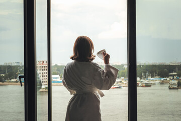 Back view of beautiful young woman in bath terry robe with cup of coffee standing and enjoying near open panoramic window of  hotel room opposite the lake.