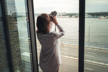 Back view of woman photographer in a transparent nightgown taking picture using camera standing near open panoramic window of apartments.