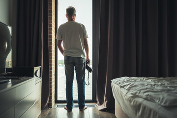 Fototapeta na wymiar Back view of middle-aged gray-haired man photographer in a taking picture using camera standing near open window of apartment or hotel room.