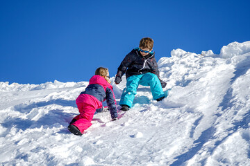 Naklejka na ściany i meble Two joyful kids playing on the snowing mountain on a winter day. Brother and sister