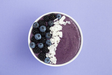 Top view of smoothies bowl with blackberries, blueberries and coconut chips on the light violet surface