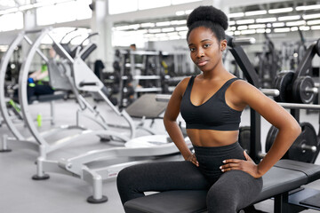 Fototapeta na wymiar portrait of black woman taking a break after workout at gym. she sits next to barbells, dumbbells, posing at camera, wearing wportswear