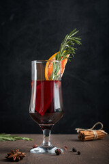 Glass of hot spiced red mulled wine usually prepared at cold winter holidays decorated with orange slices and fresh rosemary surrounded by anise star and cinnamon bunch on dark wooden table