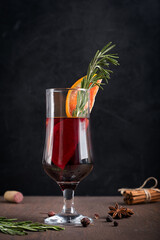 Glass of spiced hot red mulled wine usually prepared at cold winter evenings decorated with orange slices and fresh rosemary in front of cinnamon bunch and anise star on dark wooden table at kitchen
