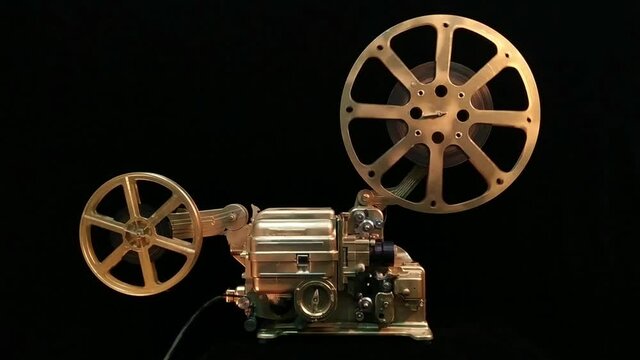 A working retro film projector of gold color in full face on a black background.
Spools of film spinning