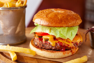 Huge burger with meat patty, green lettuce, red tomato, cucumber, cheese and sauce. Hot cheeseburger with crispy fries on wooden surface. Classic american food.