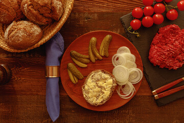 Buttered roll on wooden plate with  onion pickled cucumber vine tomato and minced meat on table