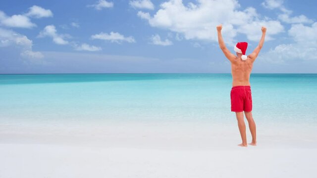Christmas CINEMAGRAPH - seamless loop: Beach man happy on travel vacation wearing santa hat happy on beach holidays happy cheering arms raised winning celebrating on sand on travel destination.