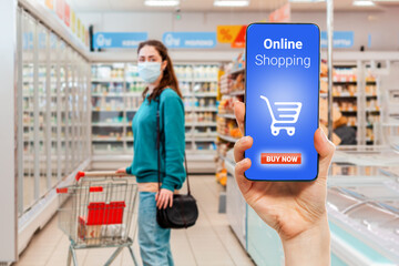 A person's hand holds a mobile phone, with a shopping basket on the screen. In the background, a woman wearing medical mask is choosing food in a supermarket, in a blur. The concept of online shopping