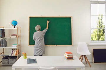 Woman teacher with gray hair writes on the blackboard in the school classroom. Back view.