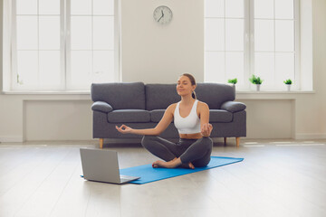 Online yoga maditation home.Attractive young girl sitting in lotus pose near laptop at home. Fit woman doing yoga, meditation or breathing exercises