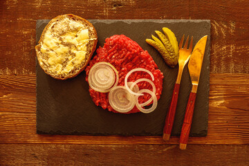 Buttered roll with minced meat sliced onion cucumber and cutlery on slate slab on wooden table