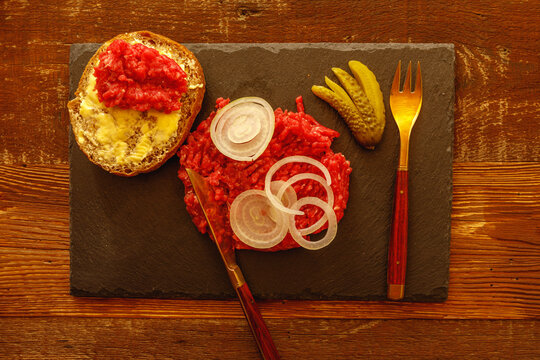Buttered roll with minced meat sliced onion cucumber and cutlery on slate slab on wooden table