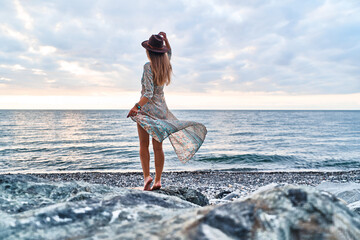 Boho chic woman in long fluttering dress and felt hat standing back on stone by the sea - Powered by Adobe