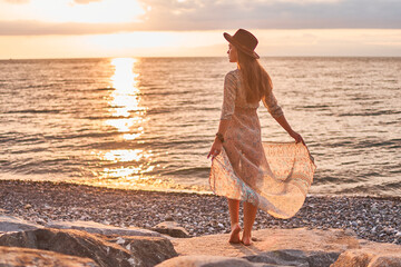 Boho chic woman in long dress and felt hat standing on stone by the sea at sunset - Powered by Adobe