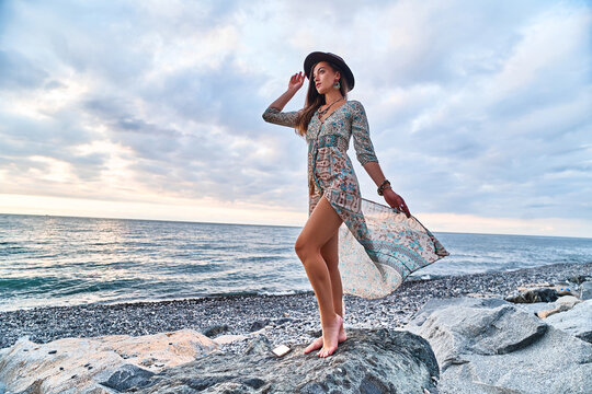Boho woman wearing jewelry with fluttering long dress and felt hat standing on a stone by the seashore