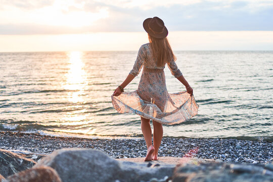 Boho chic woman in long fluttering dress and felt hat standing back on stone by the sea at sunset