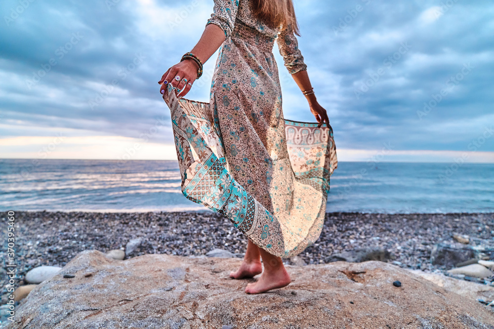 Wall mural boho woman with long waving dress standing on a stone by the seashore at sunset