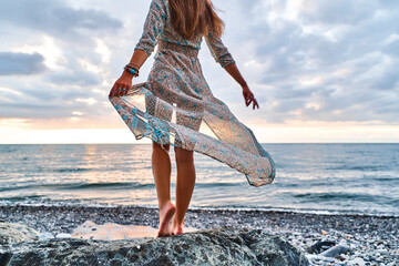 Boho woman with long legs and waving dress standing on a stone by the seashore at sunset