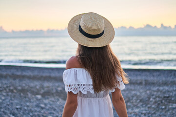 Romantic woman in a summer white dress with bare shoulders and a straw hat standing back by the...
