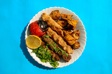 Famous traditional Arabic, Turkish, Israel food. Grilled chicken shashlik, lamb, beef kofta kebab, tomato, parsley on white plate on blue background, top view. Mixed eastern kafta kebab