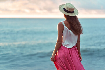 Serene dreaming romantic woman in white blouse, pink skirt and straw hat standing on the seashore and looking into a distance