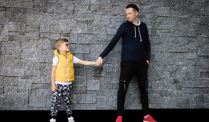 Happy father and son hold hands while looking at each other and  standing against wall. Copy space.