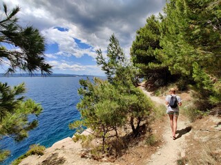 Young active feamle tourist wearing small backpack walking on coastal path among pine trees looking...
