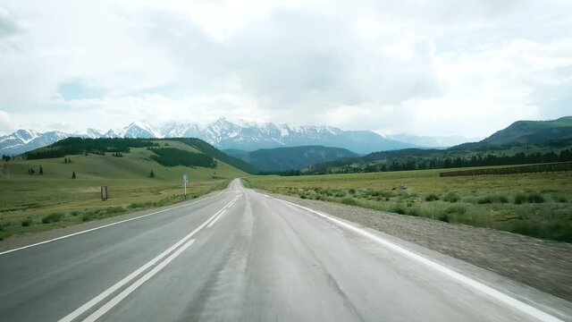 Road in the field on the background of snow-capped mountain peaks. Scene. Alps. Frontal car traveling. POV video from auto onboard mountain view on cloudy sky backa.