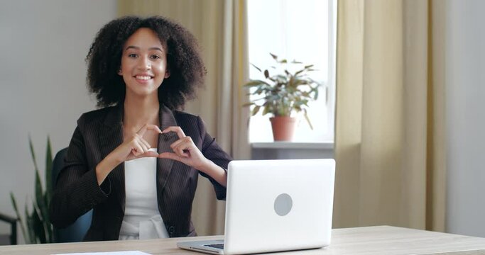 Close up friendly young African American business girl sits at office desk table with laptop, smiles toothily, folds her hands in heart sign in front of her, concept of like, hope, trust, friendship