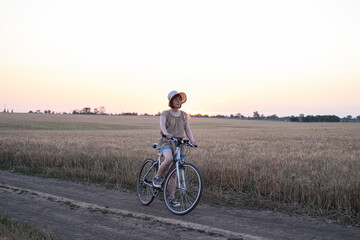 Young woman with hat ride on the bicycle in summer wheat fields