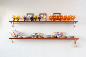 Bancada de madeira na parede repleta de taças, pratos, cumbucas, potes e objetos de vidro, porcelana e cerâmica. Decoração em parede branca