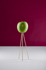 Apfel Art – grüner Apfel auf drei Holzspießen auf weißer reflektierender Oberfläche mit pinkem Hintergrund