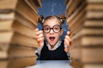 Happy smart girl in rounded glasses sitting between two piles of books and look at camera smiling.