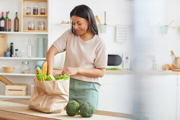 Waist up portrait of elegant mixed-race woman unpacking bags with food while standing in white...
