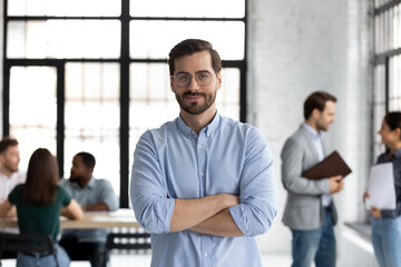 Head shot portrait smiling confident businessman wearing glasses standing in modern office room with arms crossed, diverse colleagues on background, executive boss startup founder looking at camera - obrazy, fototapety, plakaty