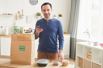 Waist up portrait of mature bearded man smiling at camera and holding smartphone while unpacking food delivery bag in kitchen interior copy space