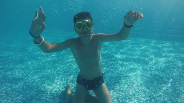 Smiling boy swimming underwater and waving one hand cheerily in a pool in slo-mo 
