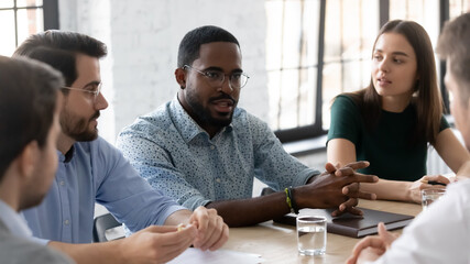 Confident African American businessman wearing glasses speaking, sharing ideas, discussing at corporate meeting, diverse colleagues listening to coach mentor speaker, sitting at table in boardroom