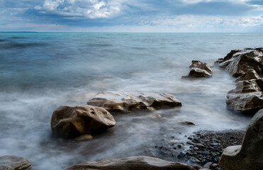 Seawaves splashing onto the sandy coast with pebbles and blue cloudy sky.