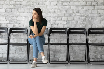 Thoughtful stressed unemployed young woman candidate waiting for job interview or hr manager employer decision, sitting on chair alone in empty office hall, feeling nervous, recruitment concept