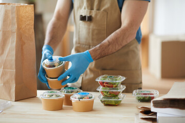 Closeup of unrecognizable worker wearing protective gloves packaging orders at wooden table in food delivery service, copy space