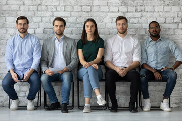 Portrait of confident diverse candidates sitting in row in queue, serious applicants business...