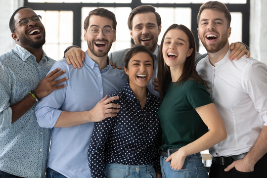 Overjoyed laughing diverse employees looking at camera together, standing in modern office room, successful business team members, colleagues celebrating corporate success, team building