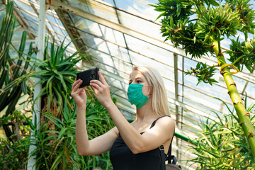 Blonde woman in face mask with mobile phone in palm greenhouse of the Botanical garden