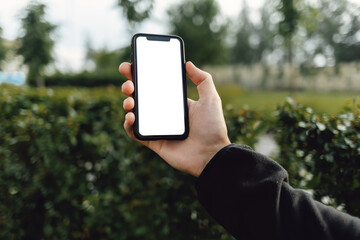 Mockup image of hand holding mobile phone with blank white screen. A man with a smartphone against the background of a city park with trees and green bushes.