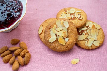 Composition in natural sunlight. Homemade gingerbread cookies with almond petals, a handful of nuts and a bowl of jam. Against the background of pink linen fabric