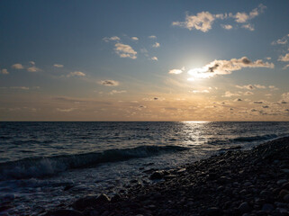 Fototapeta premium Sunset seascape, sunlight track along the water, sun in the clouds, wave runs on the pebble shore