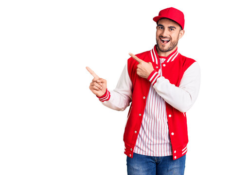 Young handsome man wearing baseball jacket and cap smiling and looking at the camera pointing with two hands and fingers to the side.